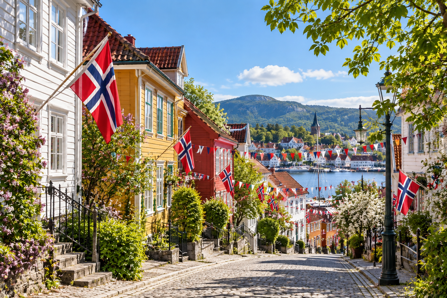 Brosteinsgate med hvite og fargerike trehus, norske flagg og vimpler, og utsikt mot sjøen på 17. mai